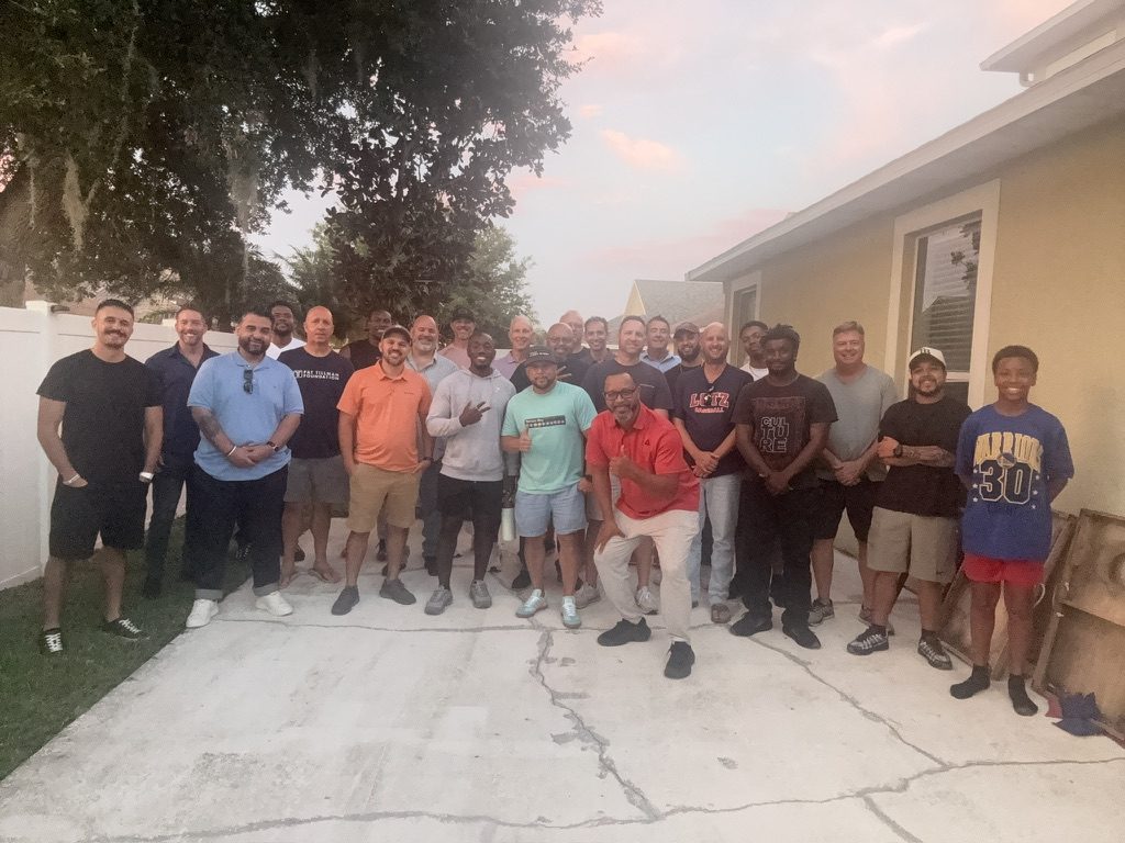 Larry Beacham kneeling in front of roughly two dozen men from the Kingdom of Disciplined Men, gathered together outdoors at sunset.